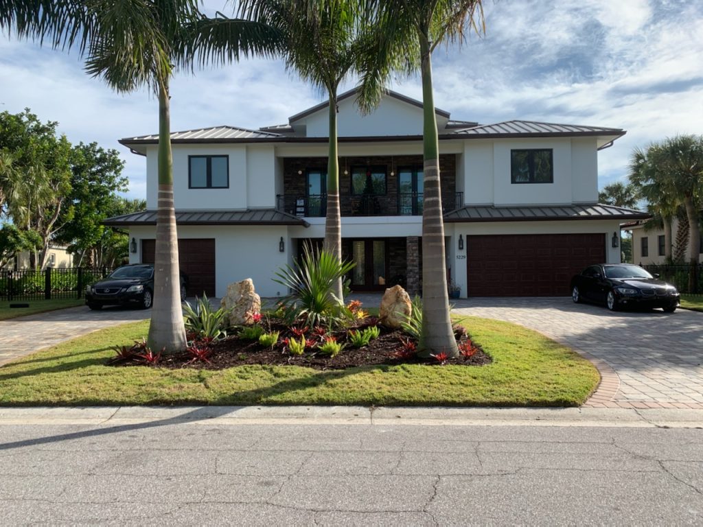 Two-story white stucco house with dual garages, tall palm trees, and landscaped front yard with two cars parked in the driveways