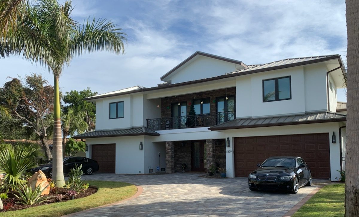 Two-story white stucco house with metal roof, balcony, paver driveway, and cars in the two-car garage