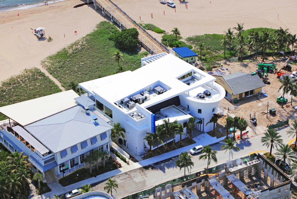 Aerial view of a beachfront building near a fishing pier, with palm trees, parking, and nearby construction sites