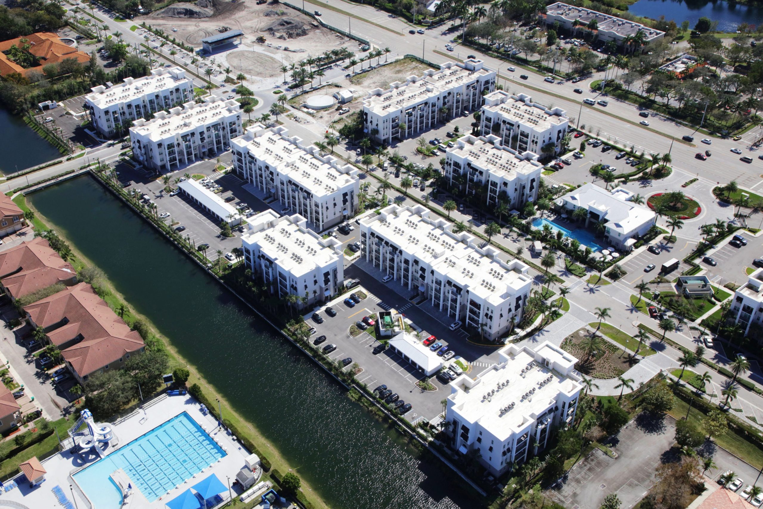 Aerial view of a white multi-building condominium complex along a canal, with pools, parking lots, and palm-lined streets