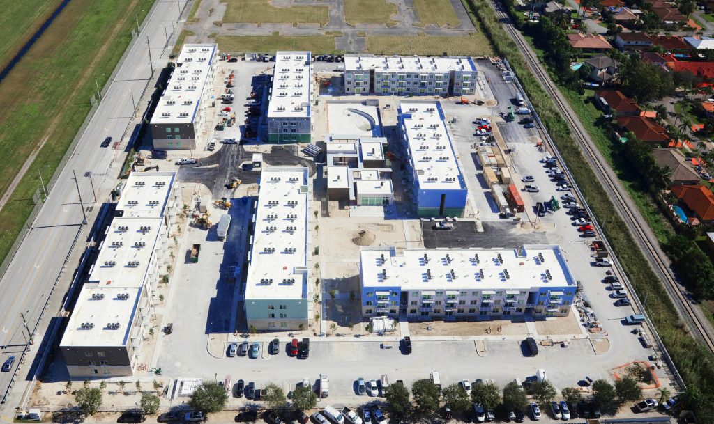 Aerial view of a multi-building apartment complex under construction, with several three-story buildings, a pool area, parking lots, and construction equipment near a residential neighborhood