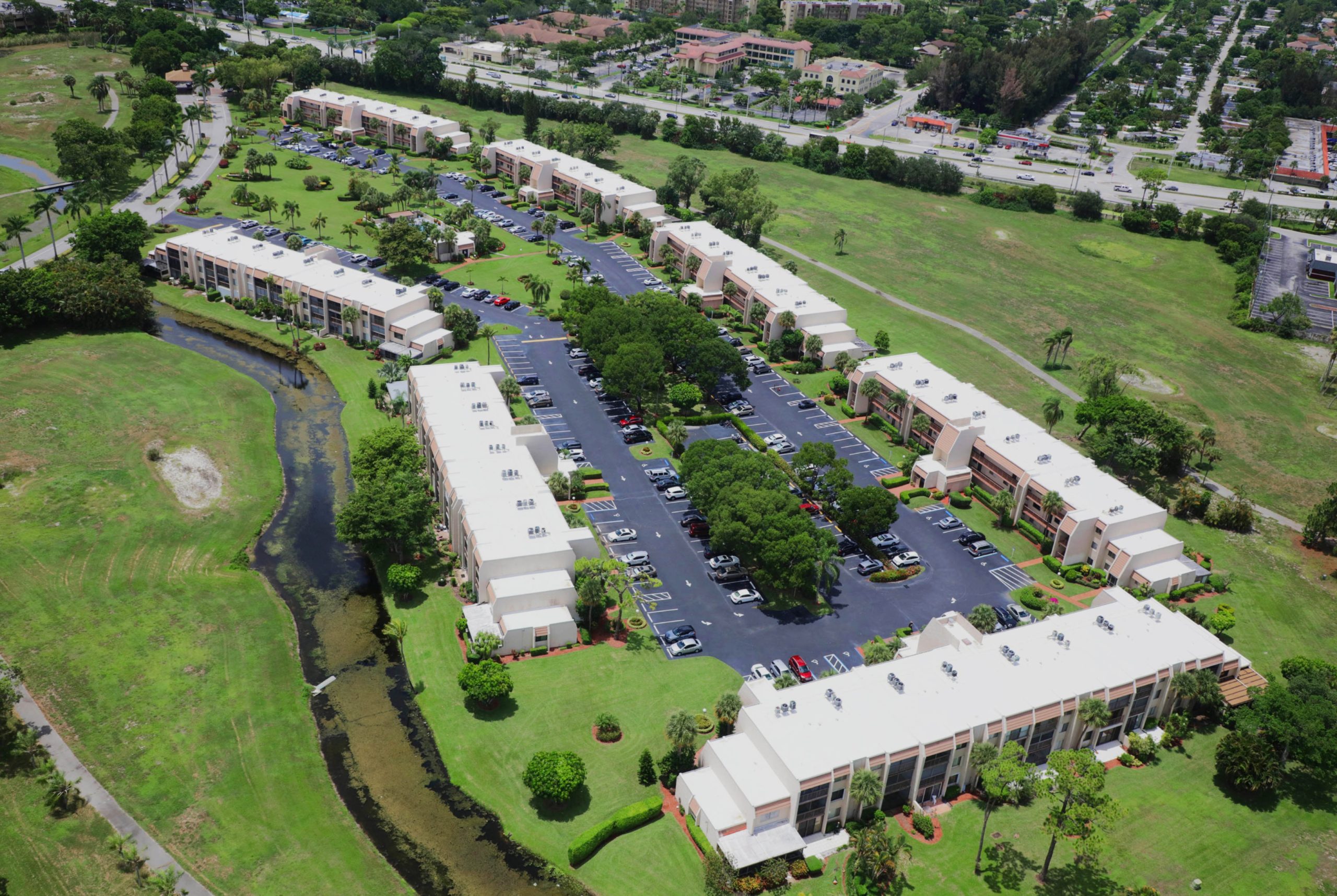 Aerial view of a condominium complex with multiple white buildings, parking lots, lawns, and a winding canal alongside the property