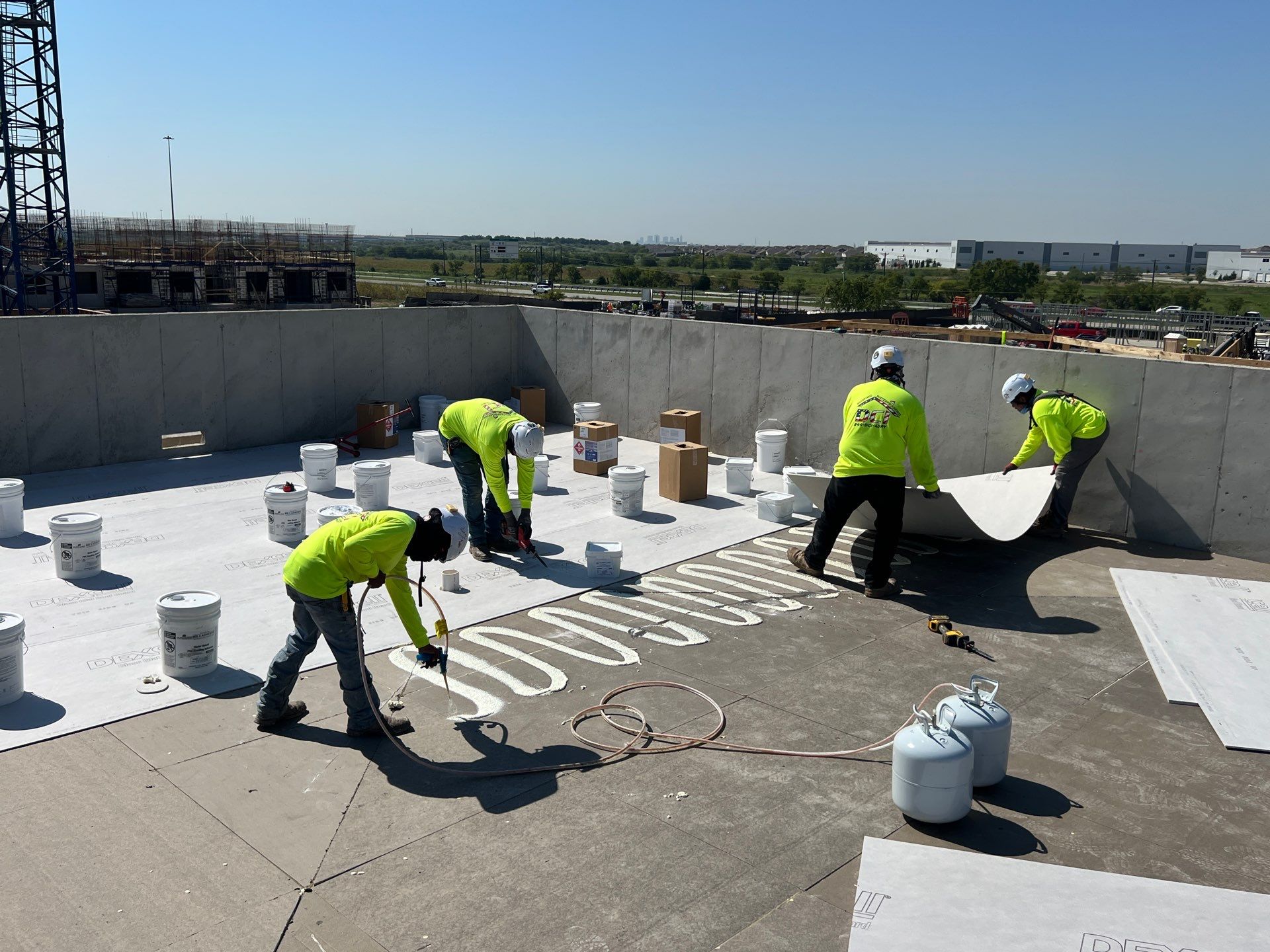 Roofing crew in yellow shirts installs insulation boards and applies adhesive on a flat rooftop under construction