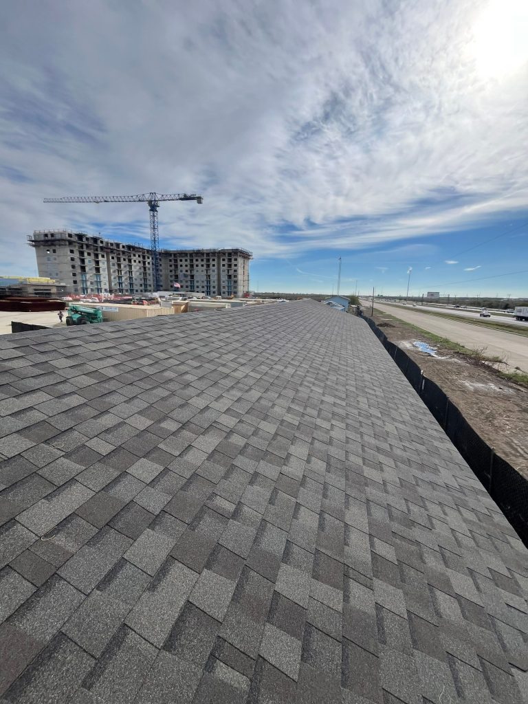 Newly installed gray asphalt shingle roof with a high-rise building under construction and highway in the background